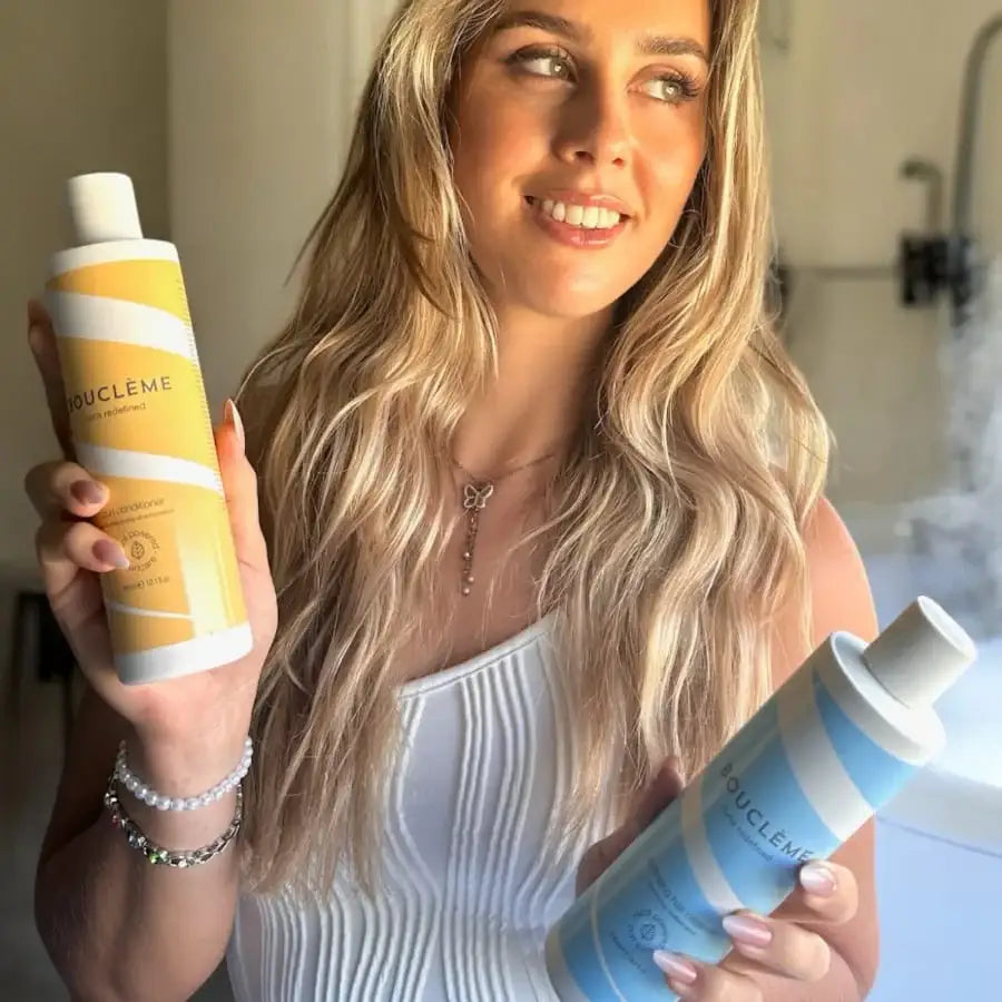 Woman with wavy blonde hair holding Bouclème curl conditioner and shampoo bottles for curly hair care