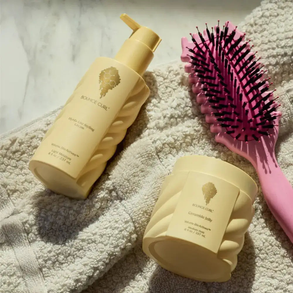 Bounce Curl styling cream and ceramide jelly with pink detangling brush on towel for curly hair care